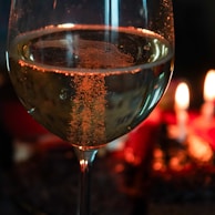 A close-up of a wine glass filled with sparkling wine, capturing the bubbles as they rise to the surface. In the background, two softly glowing candles provide a warm, intimate ambiance.