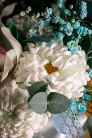 A detailed arrangement of flowers featuring predominantly white and blue tones. The central focus includes a large white chrysanthemum with a yellow center surrounded by smaller bright blue flowers and greenery, including eucalyptus leaves. The arrangement is complemented by lace and ribbon accents, creating a delicate and fresh appearance.