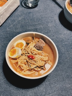 A bowl of ramen is placed on a denim blue tablecloth. The ramen contains noodles, a boiled egg cut in half with a soft yolk, slices of meat, mushrooms, and a red chili pepper in a broth.