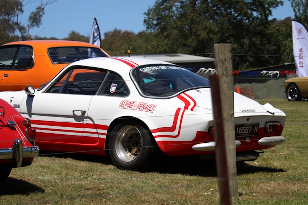 A classic Alpine Renault car with a white body and red stripes is parked on a grassy area. In the background, other vintage cars are visible, including an orange vehicle. Trees and banners can be seen surrounding the area, indicating a car show or gathering.