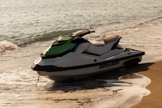 A jet ski with white and green accents rests partially on the sandy beach as gentle waves wash around it. The sea is calm with small ripples extending to the horizon.