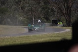 A green open-top sports car drives along a winding race track surrounded by grass and trees. In the background, several people wearing high-visibility vests are standing near a metal barrier, possibly watching the event. The scene is dust-filled and appears to be captured in a wooded area.