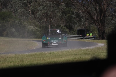 A green open-top sports car drives along a winding race track surrounded by grass and trees. In the background, several people wearing high-visibility vests are standing near a metal barrier, possibly watching the event. The scene is dust-filled and appears to be captured in a wooded area.