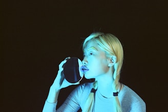 A person with light blonde hair styled in braids holds a circular mirror up to their face. The image is uniquely stylized with bold, blue lighting and accented makeup, including brightly colored eyebrows and lips. The background is dark, enhancing the contrast and focus on the subject.