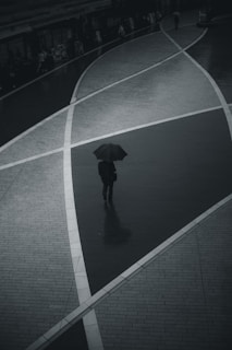 A black and white photo capturing a lone figure walking down a rainy street, umbrella in hand.