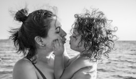 A black and white photograph of an adult and a young child by the sea. The adult is holding the child close, both facing each other. The adult has their hair tied up in a bun, and the child has curly hair. Their expressions convey a sense of tenderness and affection.