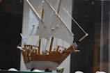 A wooden model ship with detailed sails and rigging is displayed in a glass case. The model features white sails and natural wood tones, with various ropes and pulleys intricately arranged. The background is blurred, focusing the attention on the craftsmanship of the ship model.