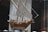A wooden model ship with detailed sails and rigging is displayed in a glass case. The model features white sails and natural wood tones, with various ropes and pulleys intricately arranged. The background is blurred, focusing the attention on the craftsmanship of the ship model.