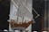 A wooden model ship with detailed sails and rigging is displayed in a glass case. The model features white sails and natural wood tones, with various ropes and pulleys intricately arranged. The background is blurred, focusing the attention on the craftsmanship of the ship model.