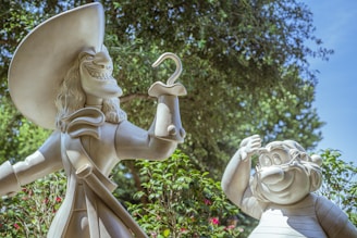 Two large sculptures of cartoon characters are set outdoors, surrounded by green foliage and a clear blue sky. One character, wearing a large hat and sporting a hook for a hand, has a mischievous grin. The other character is shorter, with a round face and an amused expression, appearing to wave.