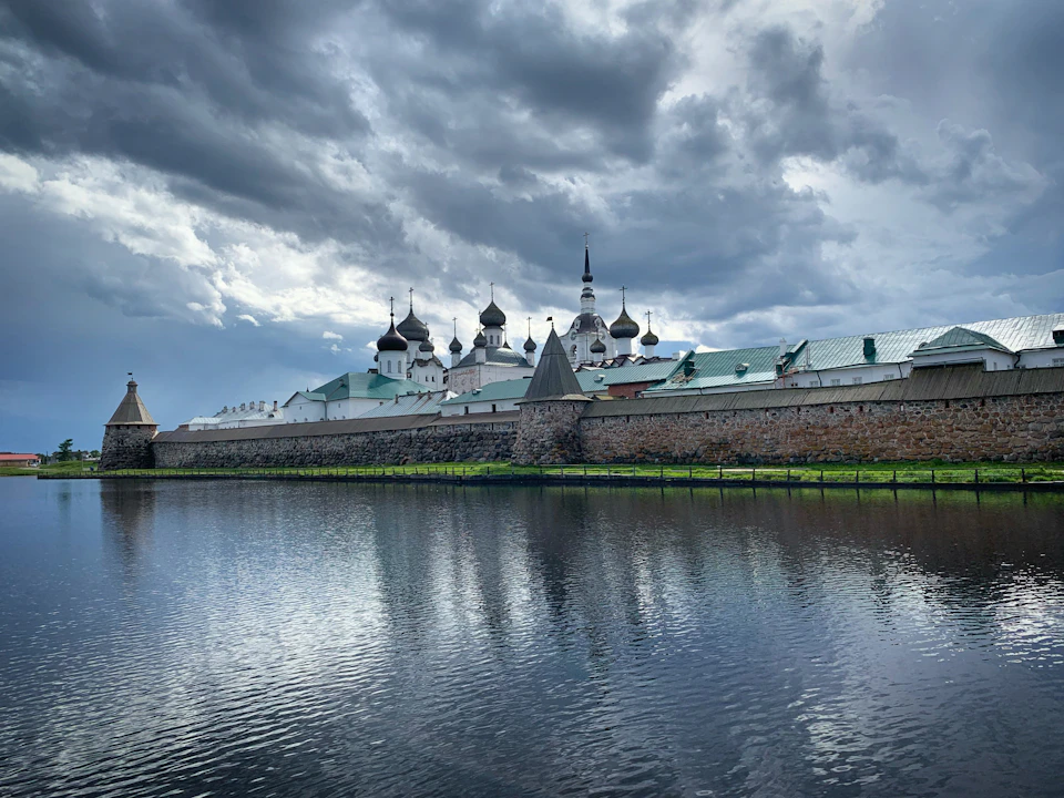 The image depicts a large, multi-tiered castle-like structure with multiple towers and domes situated on the shoreline of a lake or river. The building is constructed from stone and features intricate architectural details that showcase its historical significance. The sky above the scene is filled with clouds, creating an atmospheric backdrop for the majestic edifice.