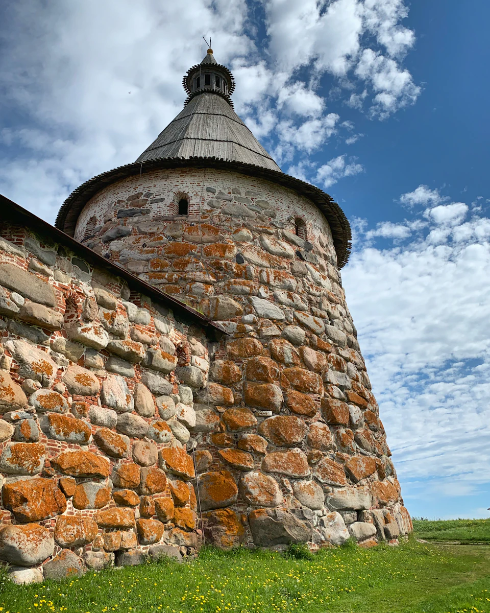 The image depicts a stone tower with a pointed roof, standing tall against a clear blue sky dotted with white clouds. The tower is constructed from large rocks and features multiple windows on its sides. The tower's position suggests it may be part of an old building or castle-like structure. In the foreground, there are patches of grass and wildflowers, adding a touch of nature to the scene.