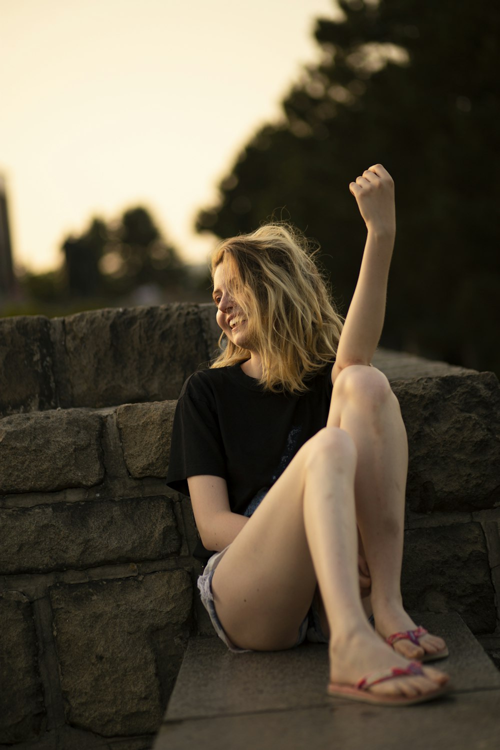 a woman sitting on a stone wall with her arms in the air