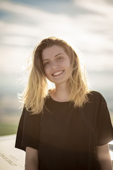 A young person with long blonde hair is smiling warmly, standing outdoors with a blurred background of a bright sky. The sunlight creates a soft halo effect around their hair, adding a serene and peaceful quality to the image.