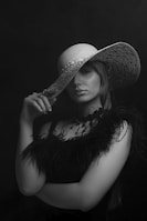 A woman poses gracefully wearing a large, stylish wide-brimmed hat and a feathered garment. Her expression is poised and confident, with a subtle hint of mystery as she holds the brim of her hat with one hand. The monochrome palette enhances the elegance and sophistication of the scene.