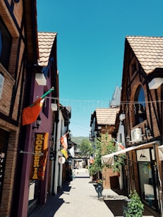 A charming narrow street lined with picturesque shops featuring wooden and brick facades. The buildings have peaked roofs with clay tiles, and several colorful flags are hanging outside the shops. In the distance, leafy green trees can be seen and the sky is clear blue, conveying a sunny day. Banners and signs add to the quaint atmosphere of this scene.