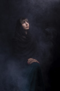 A person wearing a dark cloak is gazing upwards surrounded by a smoky, atmospheric background. Their hands are folded and resting on their lap, and the lighting creates an ethereal and mysterious ambiance.