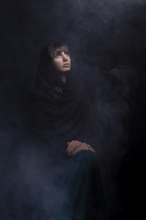 A person wearing a dark cloak is gazing upwards surrounded by a smoky, atmospheric background. Their hands are folded and resting on their lap, and the lighting creates an ethereal and mysterious ambiance.