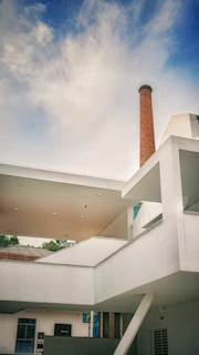 The image features a modern architectural building with a prominent white facade and geometric design. There is a tall, cylindrical brick chimney rising into a blue sky with scattered clouds. The structure has various levels, with angular shapes and flat surfaces. Trees can be seen in the background, adding a touch of greenery.