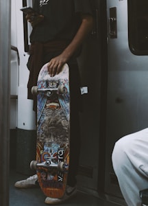 A person holds a skateboard decorated with vibrant graffiti-style artwork. The individual is standing in a subway or train with a smartphone in the other hand, dressed casually with a dark shirt and white sneakers. The atmosphere suggests urban commuting.