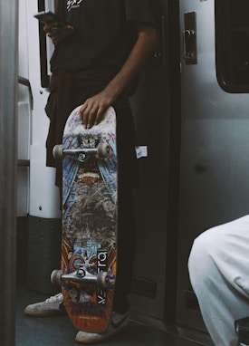 A person holds a skateboard decorated with vibrant graffiti-style artwork. The individual is standing in a subway or train with a smartphone in the other hand, dressed casually with a dark shirt and white sneakers. The atmosphere suggests urban commuting.