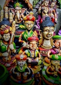 A collection of colorful, intricately designed statues depicting various deities and figures from Indian mythology. The statues feature detailed carvings and vibrant colors, with elements representing traditional musical instruments and symbolic gestures.