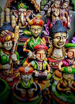 A collection of colorful, intricately designed statues depicting various deities and figures from Indian mythology. The statues feature detailed carvings and vibrant colors, with elements representing traditional musical instruments and symbolic gestures.