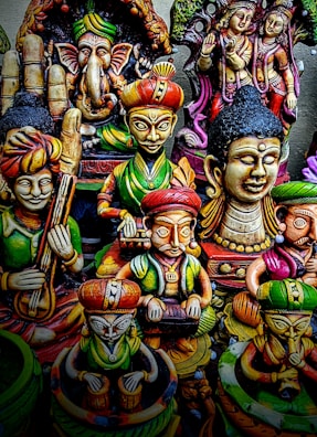 A collection of colorful, intricately designed statues depicting various deities and figures from Indian mythology. The statues feature detailed carvings and vibrant colors, with elements representing traditional musical instruments and symbolic gestures.