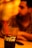 A clear glass with the Coca-Cola logo filled with a dark carbonated beverage and a red straw. In the background, a blurred figure is seated, creating a warm and cozy atmosphere with orange and yellow lighting.