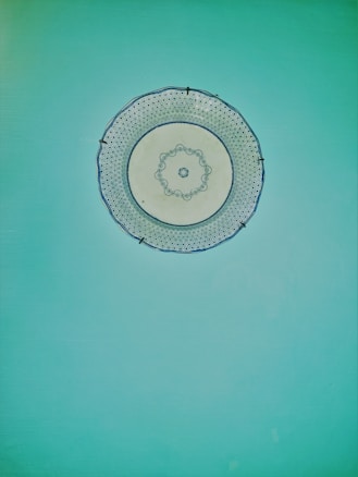 A decorative round white ceramic plate with intricate blue patterns is mounted on a bright turquoise wall. The plate features a detailed design along its edges and center, creating a contrast with the solid background.