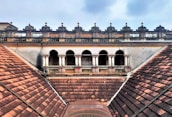 An architectural view of a historic building featuring a series of arched windows and ornate balustrades. The roof is composed of reddish-brown tiles arranged in a symmetrical pattern, which leads up to the building's decorative facade. The sky above is partially cloudy, adding contrast to the aged structure.
