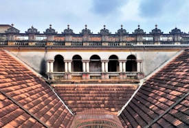 An architectural view of a historic building featuring a series of arched windows and ornate balustrades. The roof is composed of reddish-brown tiles arranged in a symmetrical pattern, which leads up to the building's decorative facade. The sky above is partially cloudy, adding contrast to the aged structure.