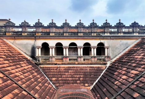 An architectural view of a historic building featuring a series of arched windows and ornate balustrades. The roof is composed of reddish-brown tiles arranged in a symmetrical pattern, which leads up to the building's decorative facade. The sky above is partially cloudy, adding contrast to the aged structure.