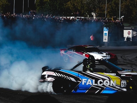 Two sports cars are drifting on a track, creating a large amount of smoke. A crowd of spectators watches from the background, while the cars maneuver through the controlled skid. The visible decals and vibrant colors on the cars add a dynamic element to the scene.