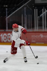 An ice hockey player in red and white gear is dynamically engaged in a game, focusing on making a shot with a hockey stick. The arena in the background is dimly lit with advertisements on the boards.