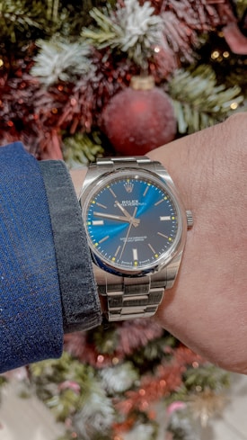 A luxury wristwatch with a stainless steel band and a blue dial is being worn by a person whose arm is visible. The watch face has silver hour markers and hands, with a recognizable brand logo. In the background, blurred decorations such as a Christmas ornament and green and red tinsel are visible, suggesting a festive or holiday setting.