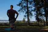 A person kneels on a grassy field, likely doing a stretch or exercise. The scene is set outdoors in a natural environment with tall pine trees casting shadows. The sky is clear with a soft blue hue. A wooden fence runs parallel to the trees, adding a rustic element to the composition.