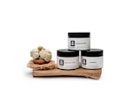 Three skincare jars labeled 'Unscented,' 'Goldflakes,' and 'Adinkra' are displayed on a wooden surface. Each jar has a minimalist white and black design. Next to them is a small bowl filled with round, off-white, cluster-like objects, possibly shea butter, sitting on a nest-like holder.