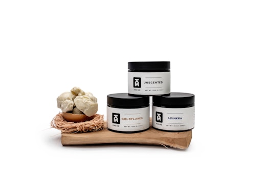Three skincare jars labeled 'Unscented,' 'Goldflakes,' and 'Adinkra' are displayed on a wooden surface. Each jar has a minimalist white and black design. Next to them is a small bowl filled with round, off-white, cluster-like objects, possibly shea butter, sitting on a nest-like holder.