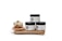Three skincare jars labeled 'Unscented,' 'Goldflakes,' and 'Adinkra' are displayed on a wooden surface. Each jar has a minimalist white and black design. Next to them is a small bowl filled with round, off-white, cluster-like objects, possibly shea butter, sitting on a nest-like holder.