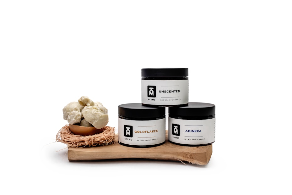 Three skincare jars labeled 'Unscented,' 'Goldflakes,' and 'Adinkra' are displayed on a wooden surface. Each jar has a minimalist white and black design. Next to them is a small bowl filled with round, off-white, cluster-like objects, possibly shea butter, sitting on a nest-like holder.