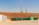 A row of green waste bins is aligned on the edge of a paved area next to a grassy slope. Above the slope, a tall communication tower is visible against a clear blue sky. A small white building with ventilation gratings is situated to the left.