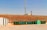 A row of green waste bins is aligned on the edge of a paved area next to a grassy slope. Above the slope, a tall communication tower is visible against a clear blue sky. A small white building with ventilation gratings is situated to the left.