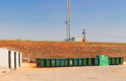 A row of green waste bins is aligned on the edge of a paved area next to a grassy slope. Above the slope, a tall communication tower is visible against a clear blue sky. A small white building with ventilation gratings is situated to the left.