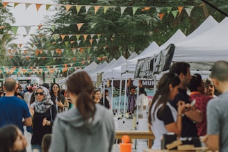 A bustling outdoor market scene with numerous people casually strolling among white canopied stalls. Colorful triangular flags are strung above the walkway, creating a festive atmosphere. Some individuals are interacting at the stalls, while others are walking through the crowd. Trees provide a green backdrop to the scene.