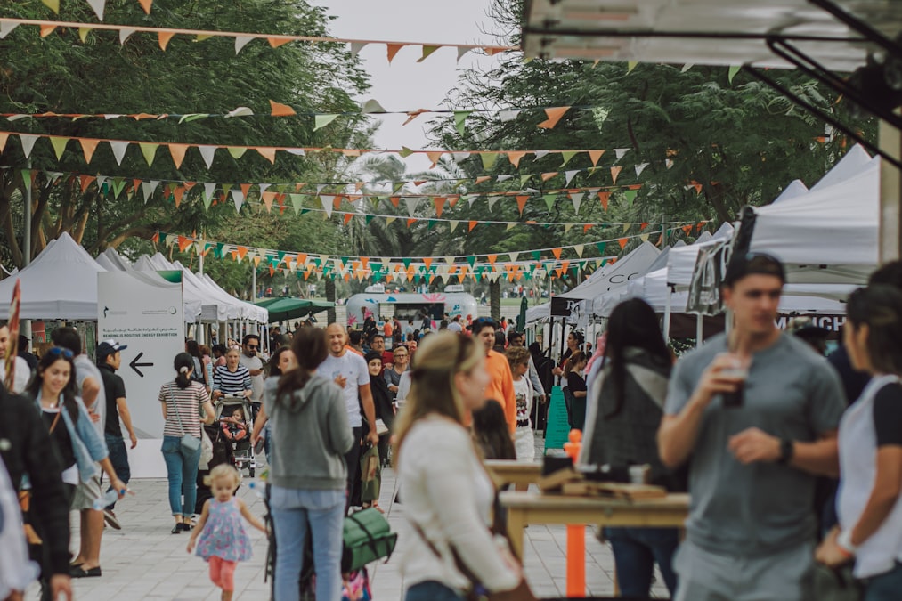 An outdoor festival or market with bunting flags and a crowd browsing white-tent vendors
