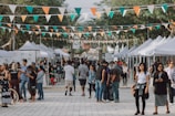 A bustling outdoor market lined with white tents, where people are engaged in shopping and socializing. Brightly colored triangular pennant flags hang overhead, adding a festive atmosphere. The crowd, consisting of various individuals, walks along the wide pathway flanked by tall trees and vendor stalls.