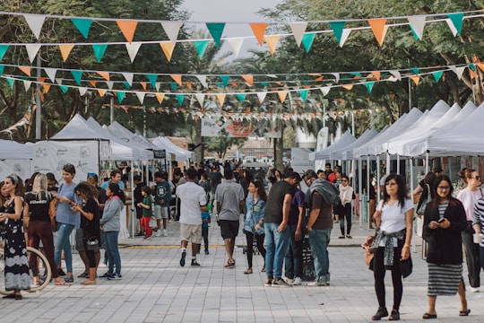 A bustling outdoor market lined with white tents, where people are engaged in shopping and socializing. Brightly colored triangular pennant flags hang overhead, adding a festive atmosphere. The crowd, consisting of various individuals, walks along the wide pathway flanked by tall trees and vendor stalls.
