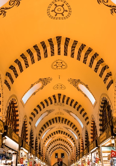 The image features an indoor market or bazaar with an ornate, arched ceiling. The ceiling is decorated with intricate patterns and designs in black against a warm yellow background. The architecture includes large arches with windows that allow natural light to illuminate the space. Stalls lined with goods and signage are visible along both sides of the corridor.