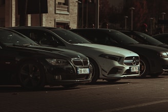 A row of parked cars under dim lighting, featuring prominently visible BMW and Mercedes models. The background includes a building with windows and partially visible pedestrians.